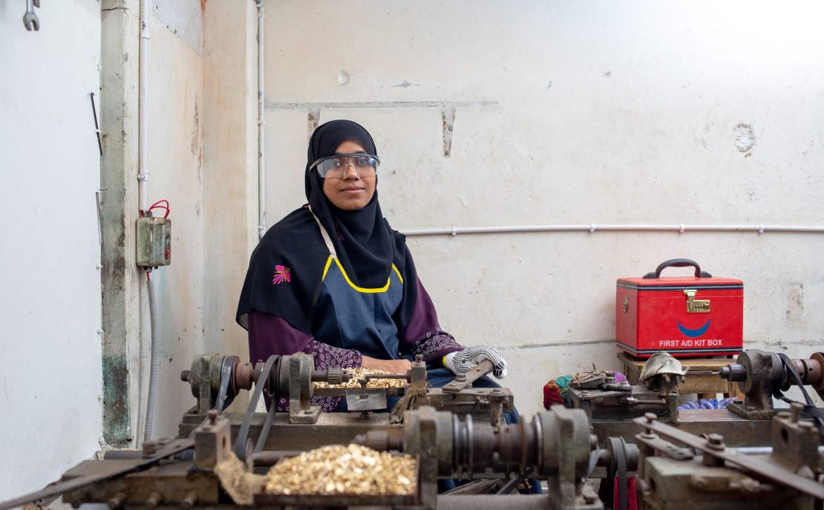 Shabiha Akter Pinky stands in her electric switch and socket manufacturing workshop in Kadamtoli, Dhaka, with a rooftop solar panel installation and workers assembling products in the background.
