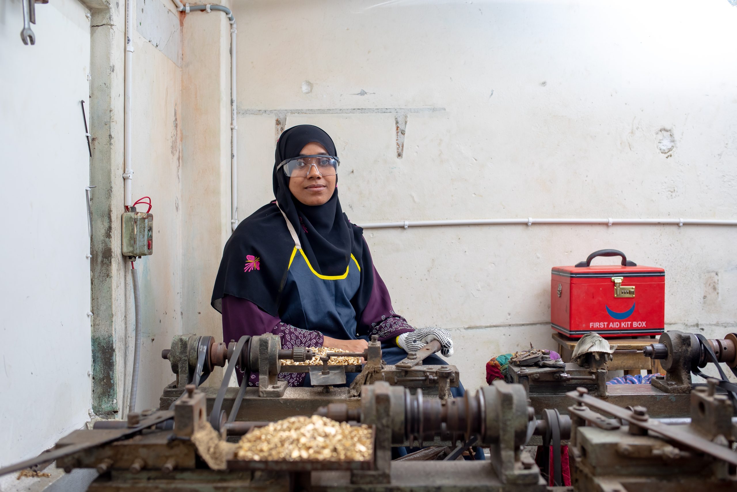Shabiha Akter Pinky stands in her electric switch and socket manufacturing workshop in Kadamtoli, Dhaka, with a rooftop solar panel installation and workers assembling products in the background.